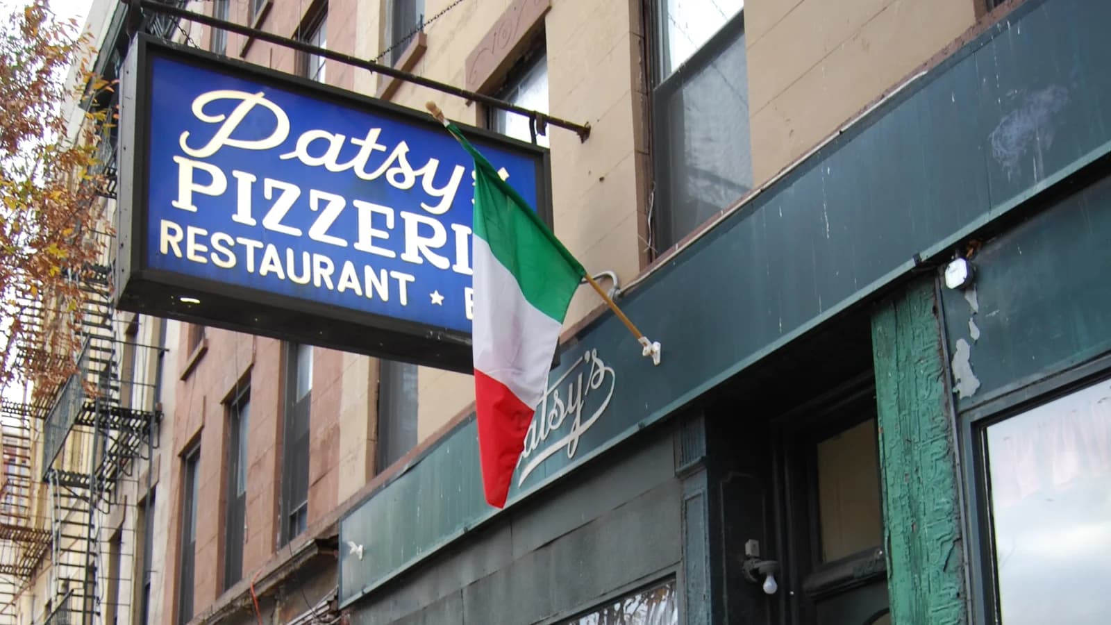 The blue Patsy's Pizzeria Restaurant sign hanging over a dark green storefront, with an Italian flag beside it and fire escapes on the brick buildings behind