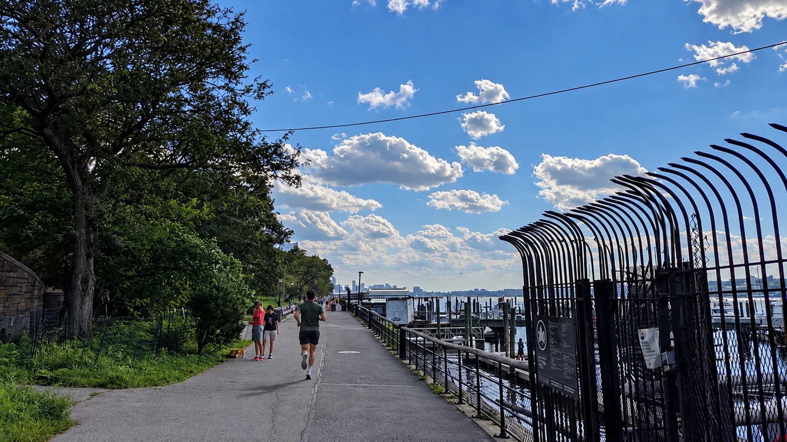 Runner in a green tee on the Hudson River Greenway in Riverside Park, with a curved black fence and the marina at right