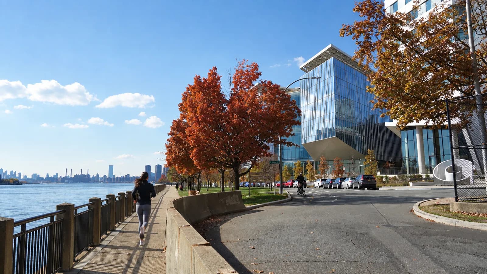 Woman in a black jacket running Roosevelt Island's East Promenade past red fall trees and the Cornell Tech buildings, with the East River at left