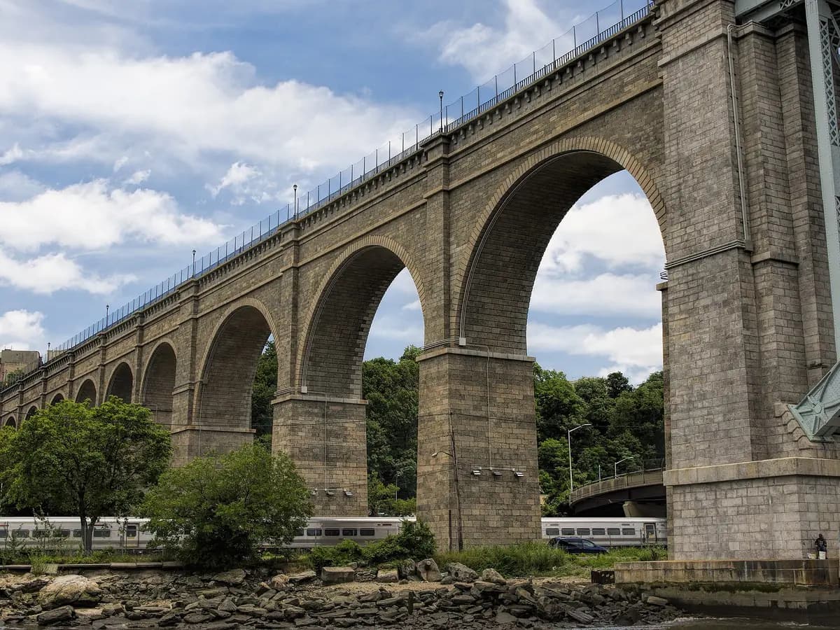 Massive stone arches of the High Bridge over the Harlem River shore, with the walkway railing running along the top under a blue sky