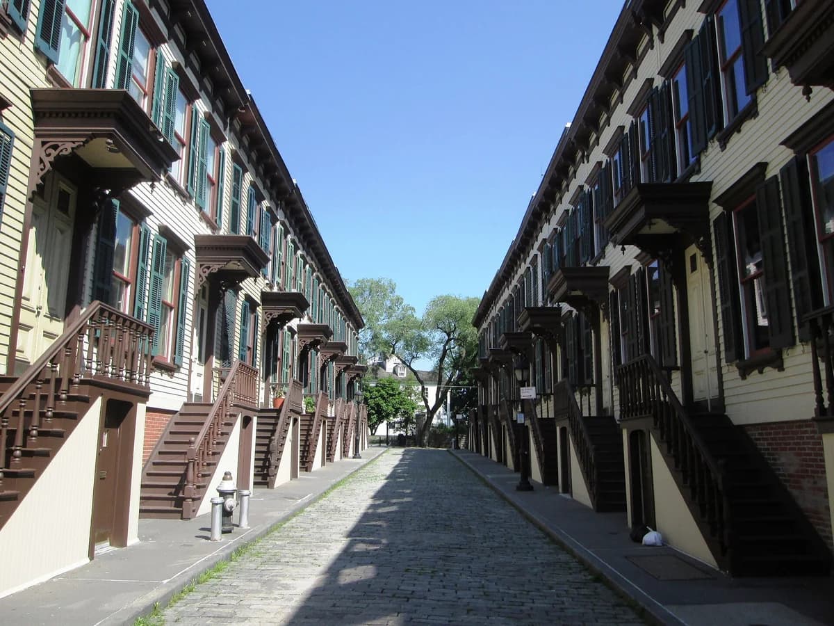 Cobbled Sylvan Terrace lined on both sides by matching yellow wooden row houses with dark shutters and steep wooden stoops