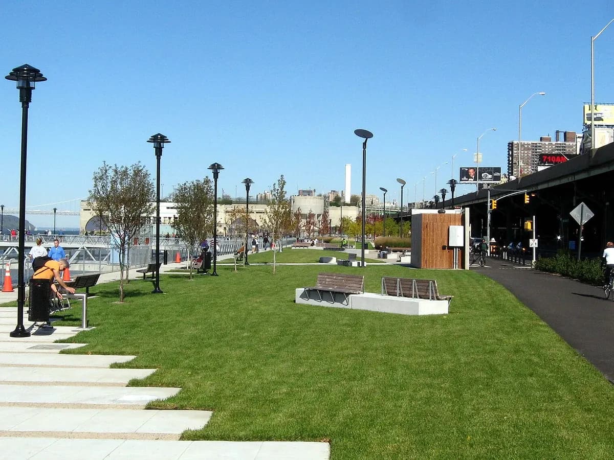 West Harlem Piers Park lawn and paved walk with tall lamp posts beside the Hudson River on a sunny day, with the Riverside Drive viaduct at the right edge