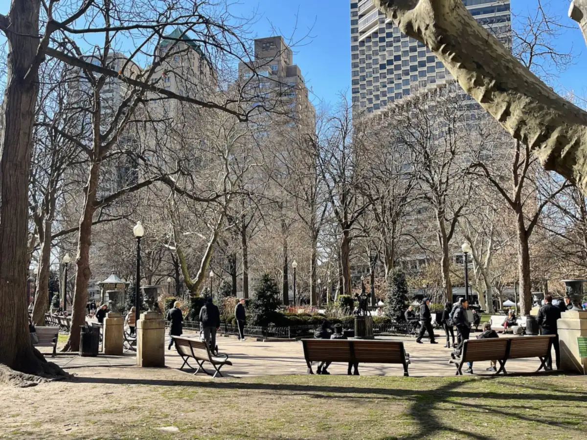 Bare winter trees and wooden benches around the paths of Rittenhouse Square, with tall apartment buildings rising beyond the lawn