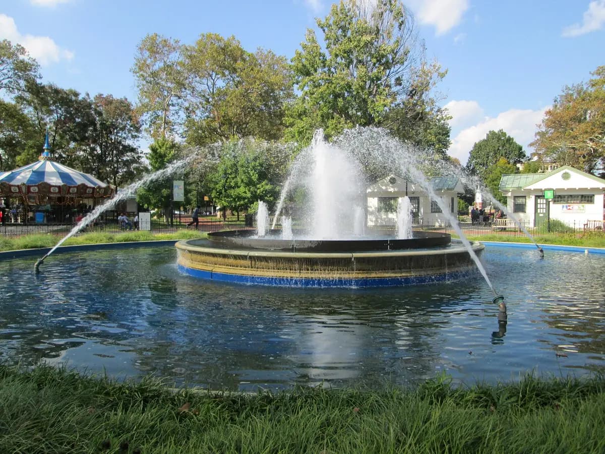 Restored marble fountain spraying arcs of water in a round basin at Franklin Square, with the carousel and trees behind
