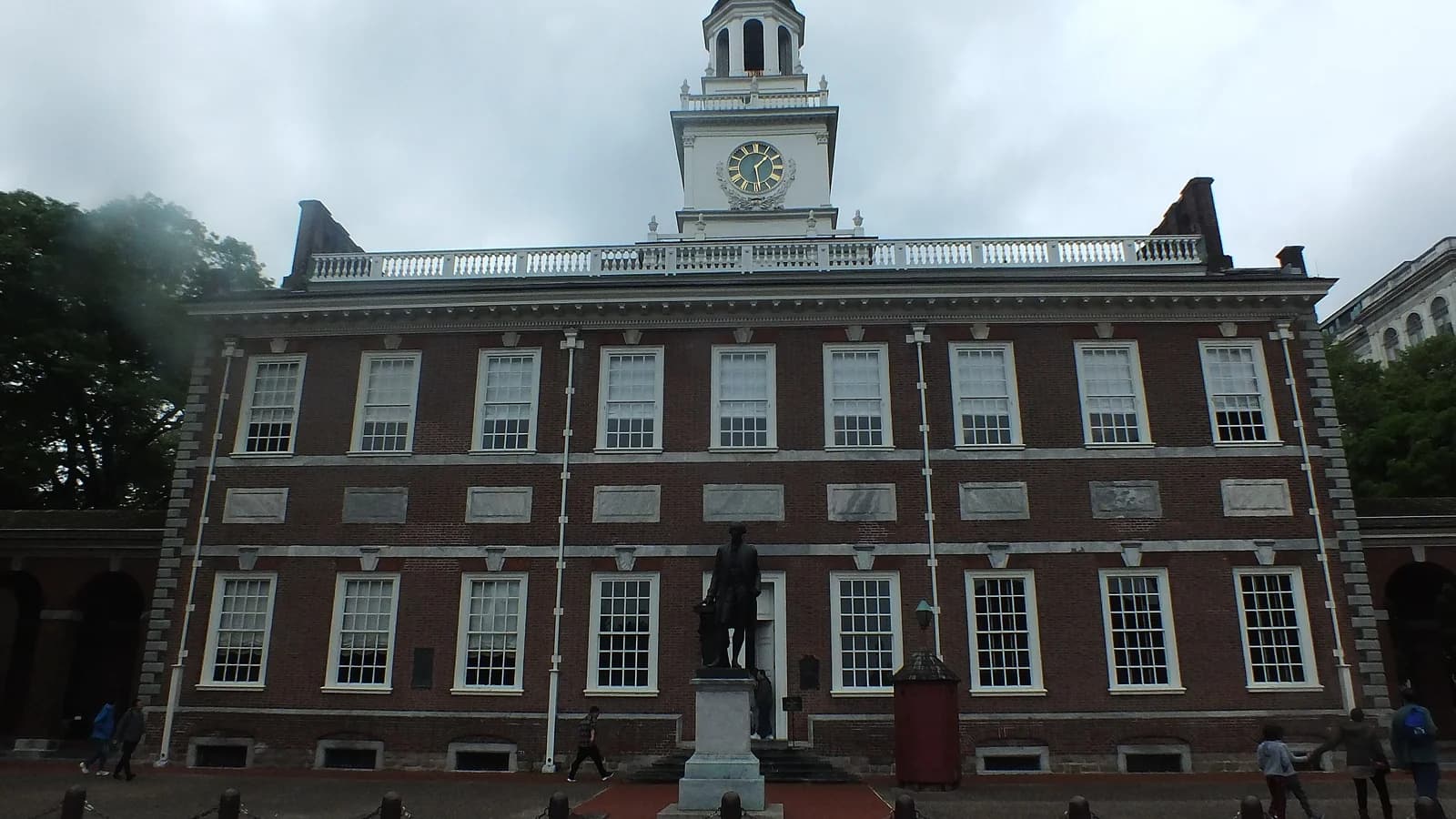 Red brick Independence Hall and its white clock tower under a cloudy sky, with a statue out front