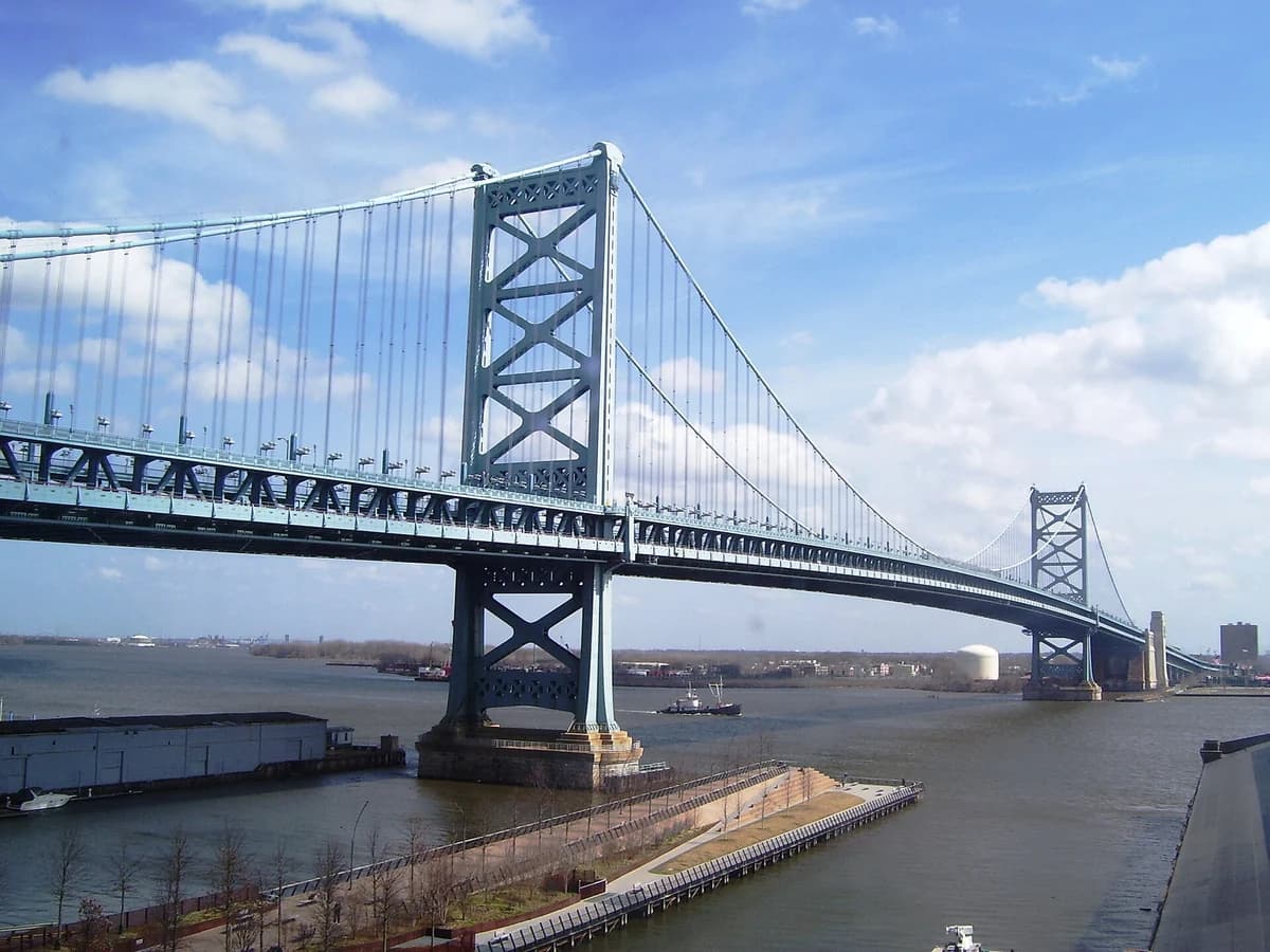 Blue steel towers and suspension cables of the Benjamin Franklin Bridge spanning the Delaware, with the Race Street Pier's landscaped edge below