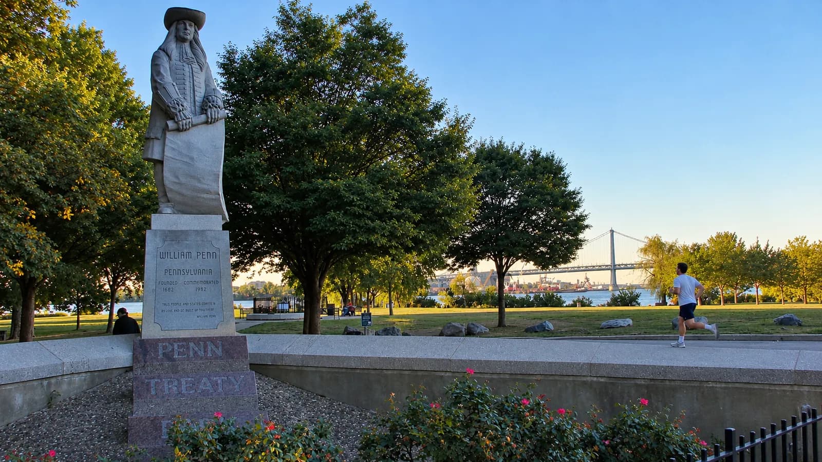 Runner in a white tee passing the William Penn statue in Penn Treaty Park, with the Delaware River and a bridge behind