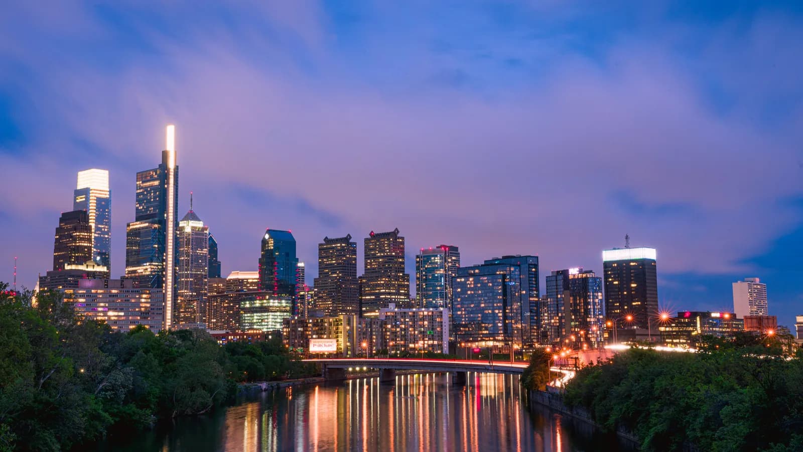Philadelphia skyline lit up at dusk and reflected in the Schuylkill River, with a bridge and green riverbanks