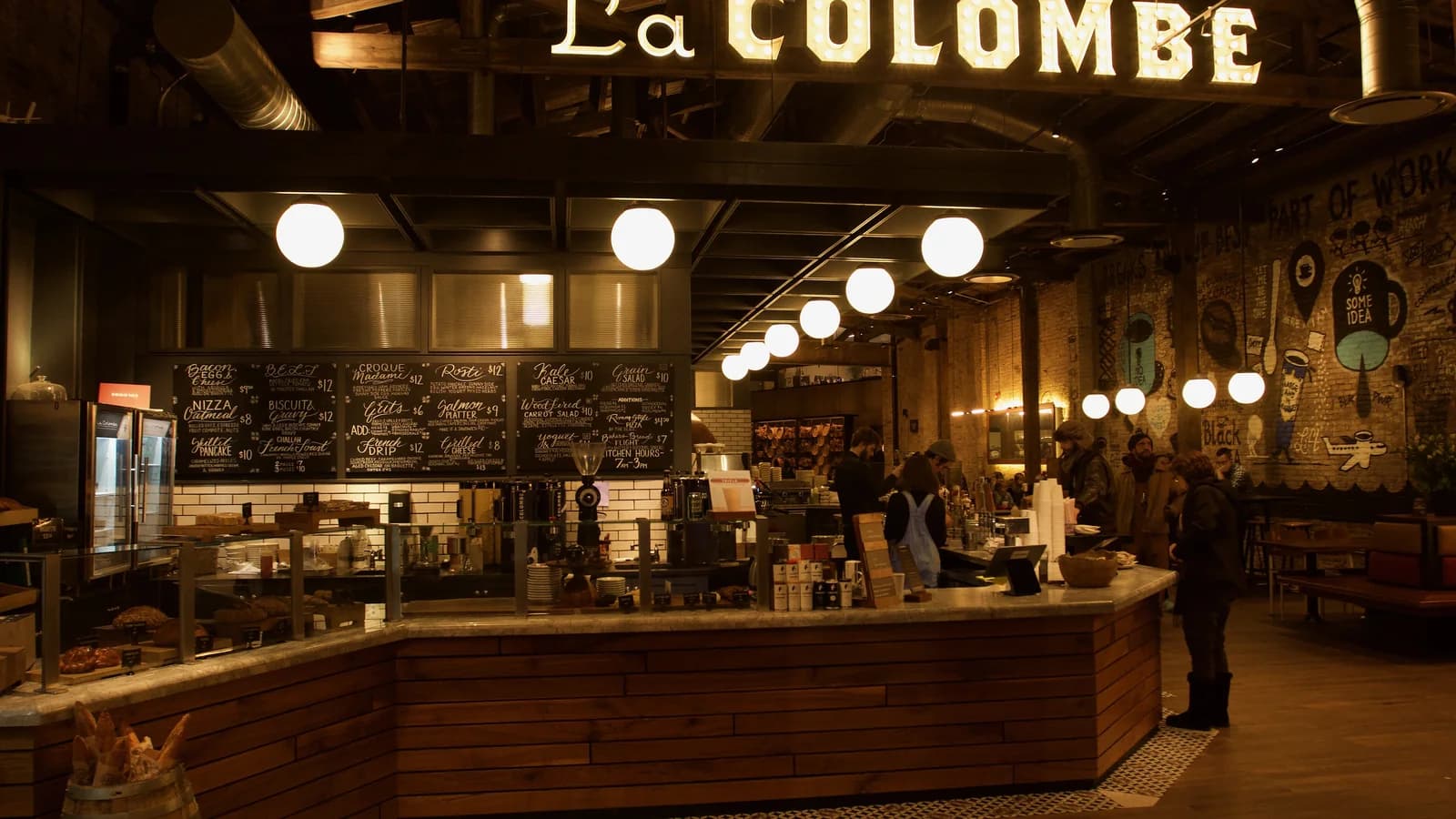 Warm globe lights over the long wooden coffee bar under the La Colombe sign at its Fishtown roastery