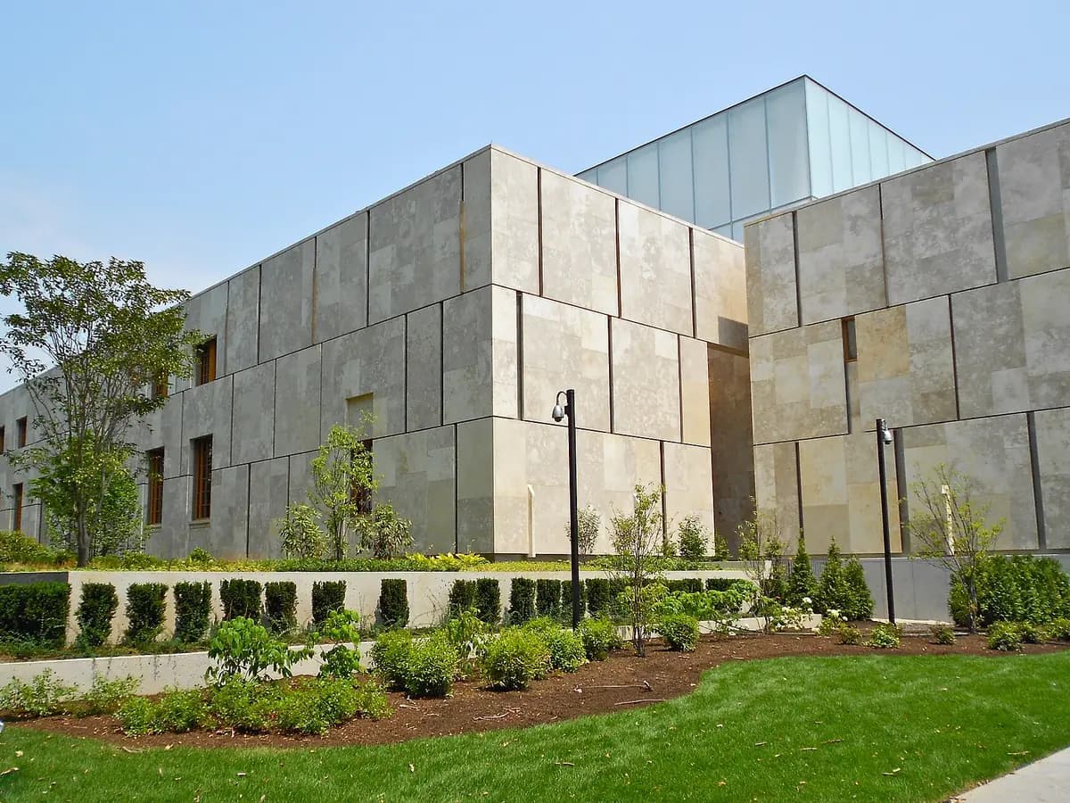 Pale limestone walls of the Barnes Foundation with a glass topped upper box, behind trees, a low wall and planted gardens on a sunny day