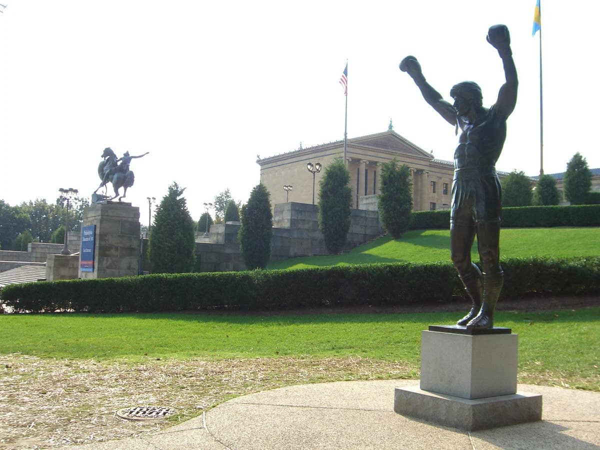 Bronze Rocky statue with both arms raised on a stone pedestal, with the Philadelphia Museum of Art columns and a mounted horseman statue on the lawn behind