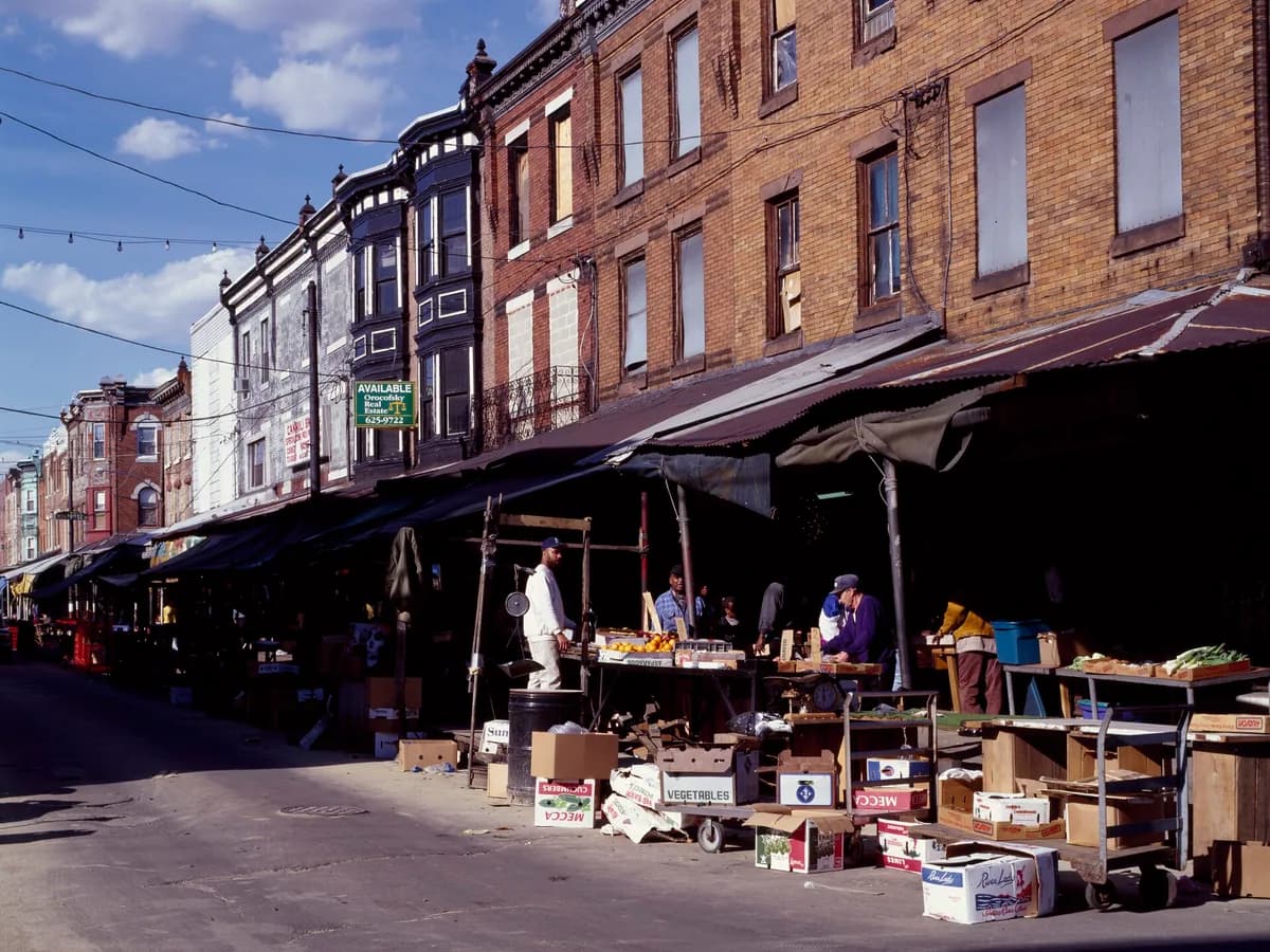 Metal awnings over produce stands along the Italian Market, with brick storefronts above and crates of vegetables stacked on the sidewalk