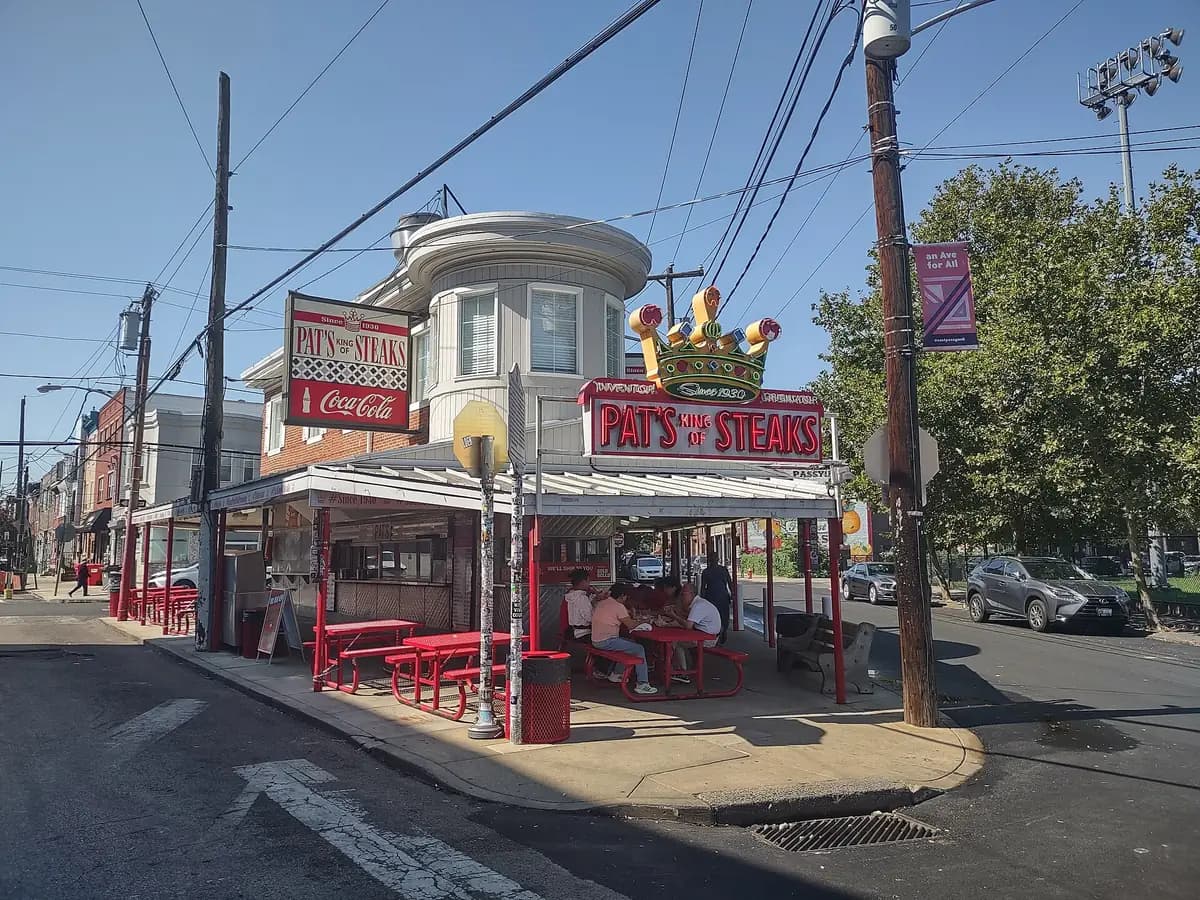 Pat's King of Steaks walk up stand on the corner, with its crown sign, red picnic tables and diners under the awning on a sunny day