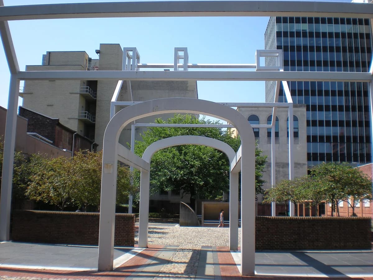 White steel ghost structure of arches and beams outlining Benjamin Franklin's lost house, framing a brick courtyard and trees at Franklin Court