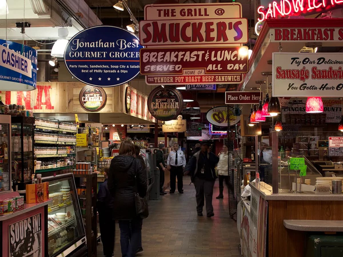 Busy aisle inside Reading Terminal Market lined with lunch counters under bright neon and painted signs, with shoppers walking between the stalls