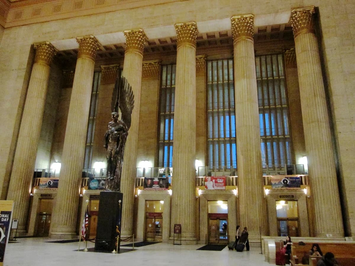 Bronze winged angel statue on a dark pedestal in the main hall of 30th Street Station, framed by tall fluted columns, high windows and a coffered ceiling