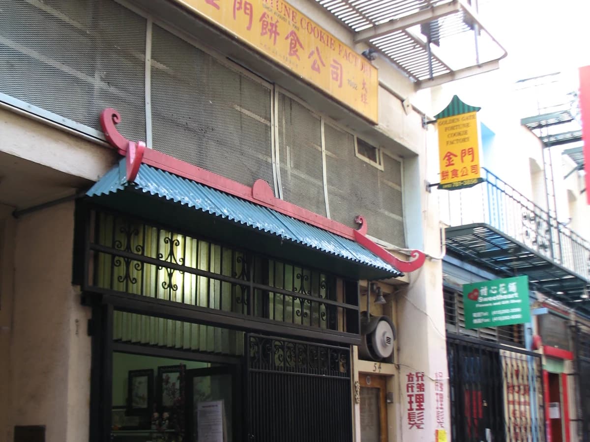 The narrow storefront of the Golden Gate Fortune Cookie Factory in Ross Alley, with a red-trimmed green awning, iron grilles and a yellow hanging sign