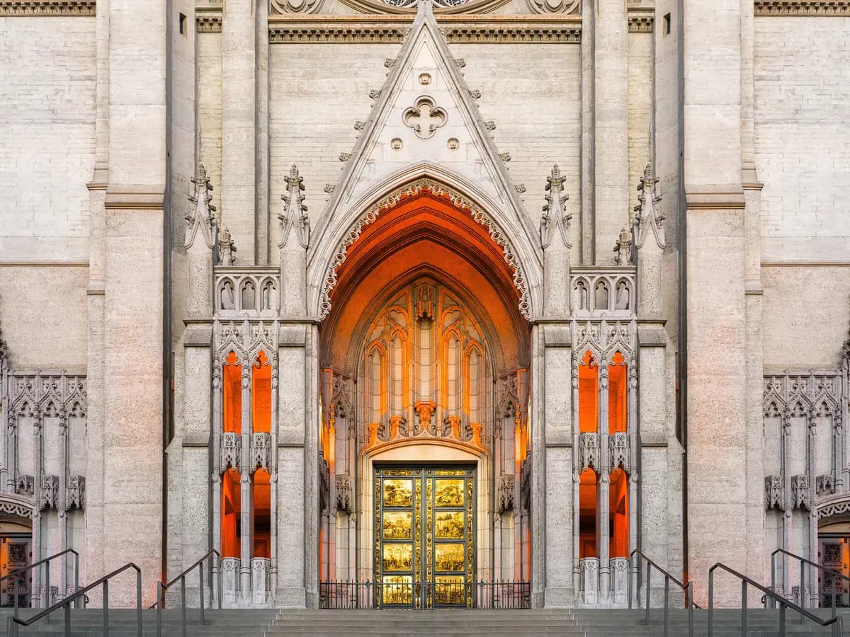 Grace Cathedral's pale Gothic central portal at dusk, with the gilded Gates of Paradise doors under a pointed arch glowing orange, flanked by pinnacles and stairs