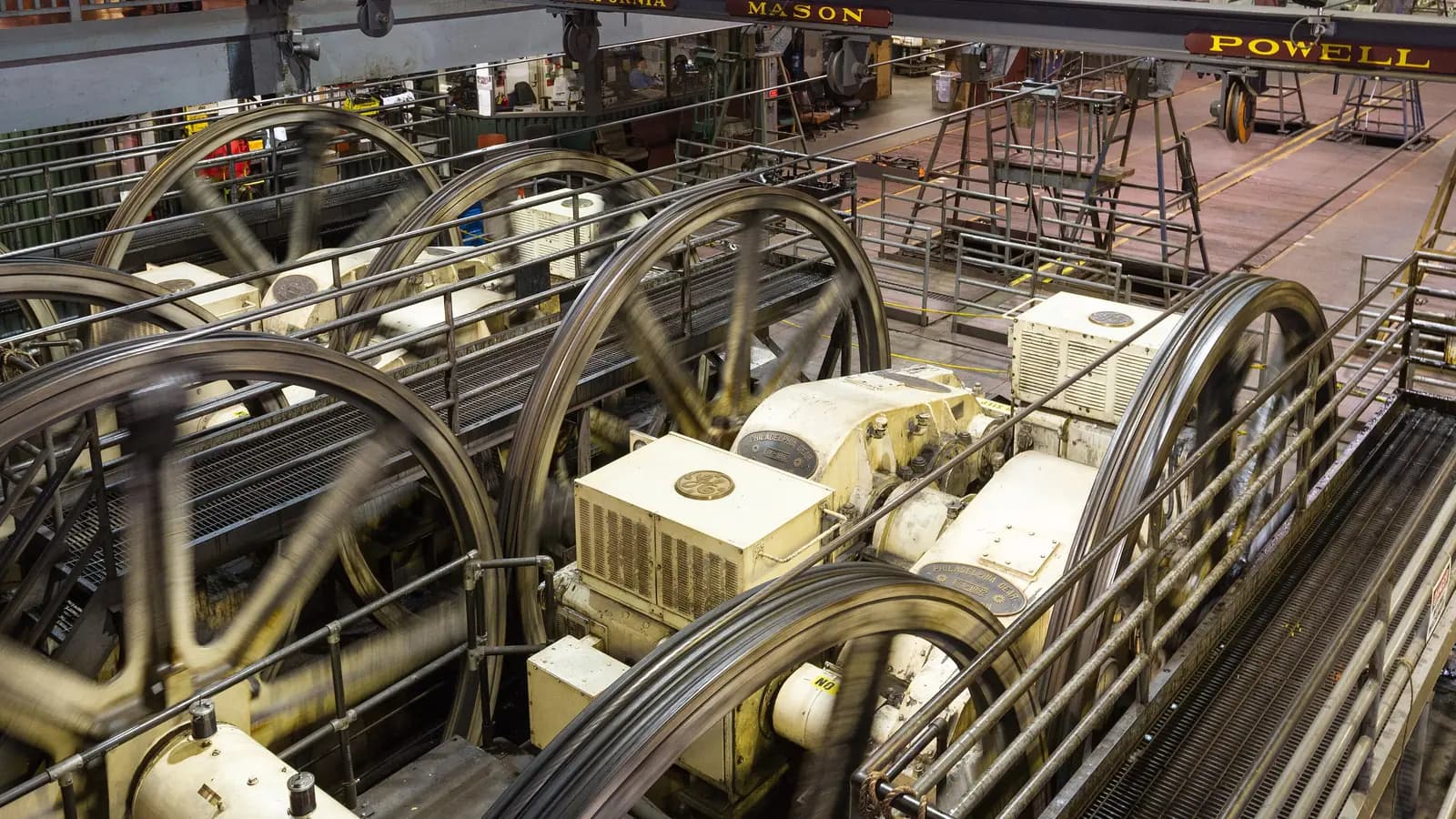 Giant winding wheels spinning as they pull the cables inside the Cable Car Museum powerhouse, under signs for the Powell, Mason and California lines