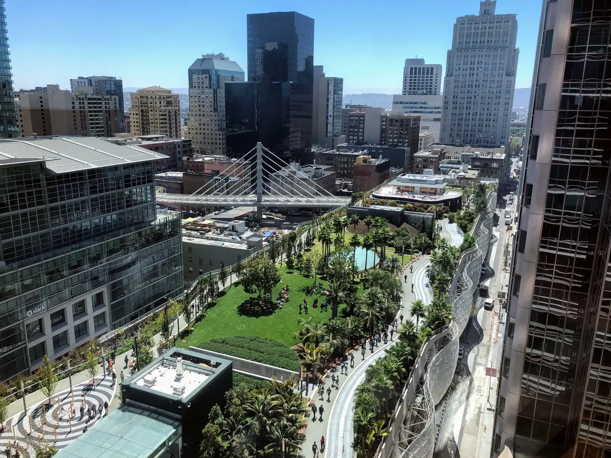 Salesforce Park seen from above: a long rooftop garden with a central lawn, palm groves and a curving walking path, ringed by office towers and a white cable-stayed bus bridge
