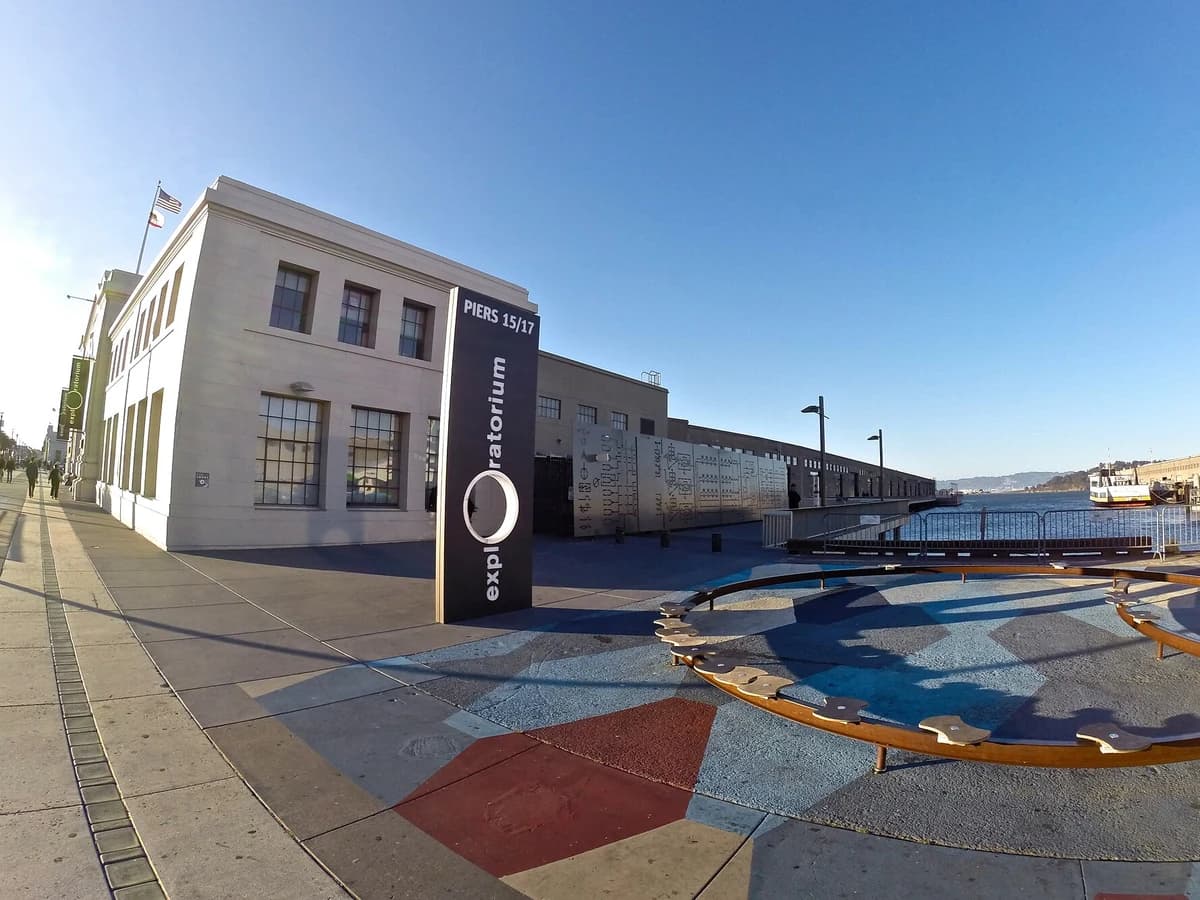 The Exploratorium's Piers 15/17 sign beside the pale pier building on the Embarcadero, with a paved plaza, curved benches and the bay beyond under a clear sky