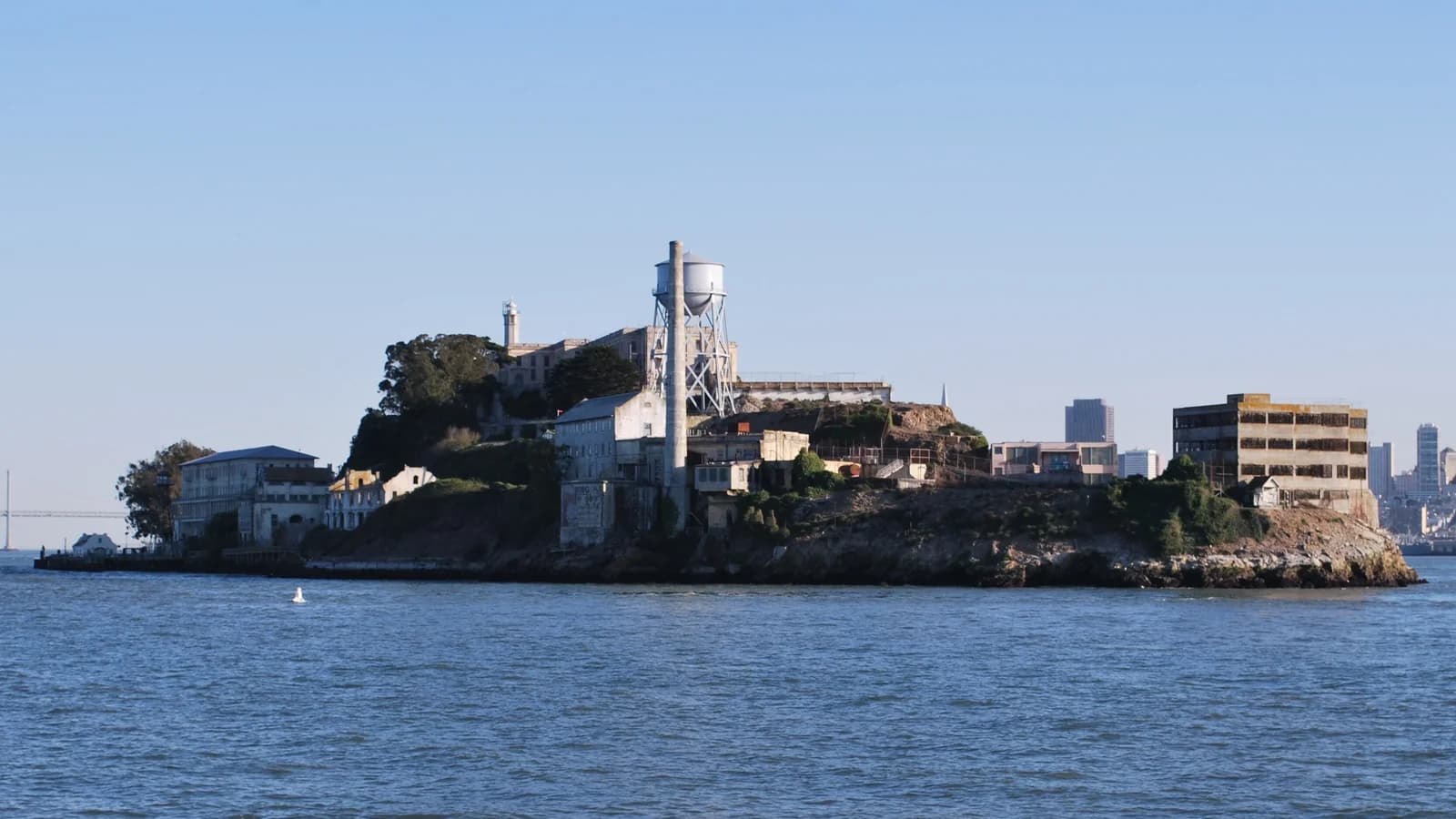 Alcatraz Island seen from the water, with the lighthouse, cellhouse and water tower on the rock and the downtown skyline in the distance