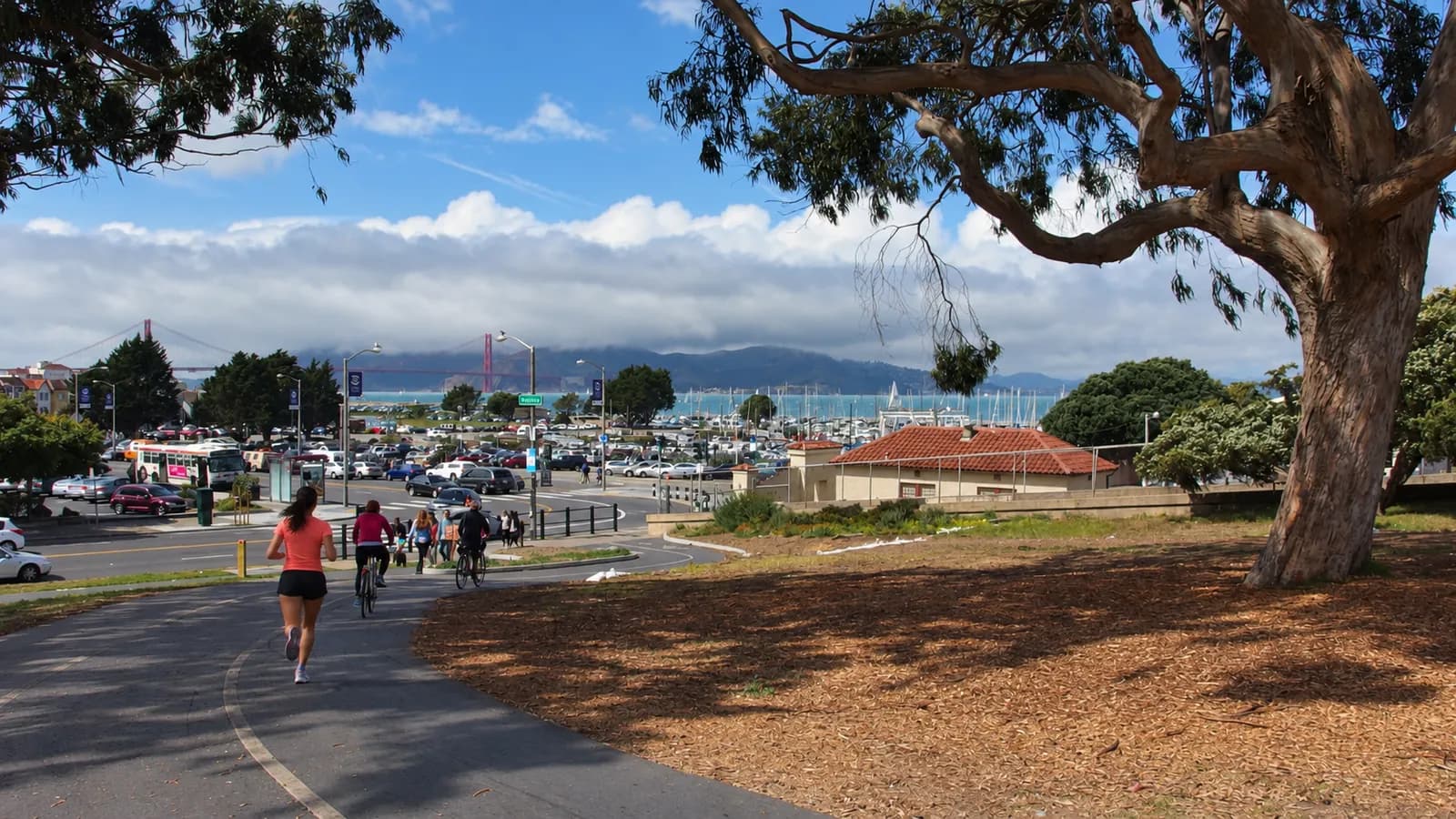 A runner in a coral top heads down a curving path at Fort Mason under a eucalyptus tree, with Marina boats and the Golden Gate Bridge in low clouds behind