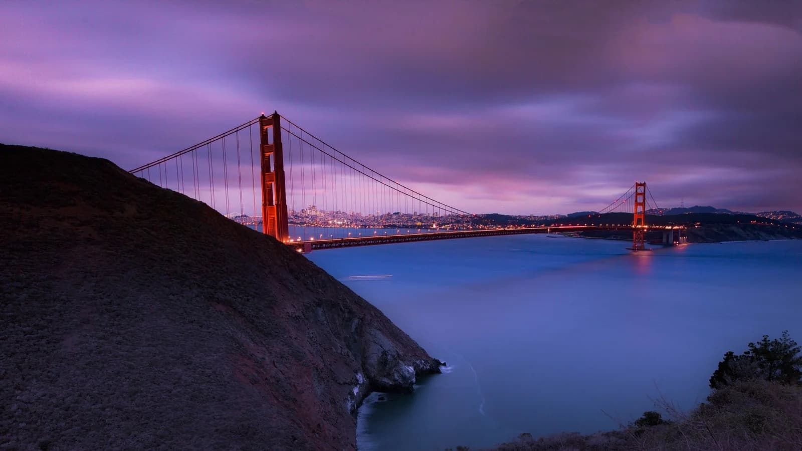 The Golden Gate Bridge's red towers lit at dusk under purple clouds, with a dark hillside in front and the lights of San Francisco on the hills across the water