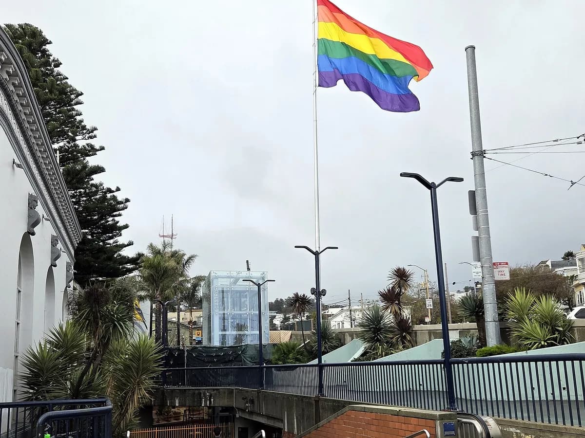 A large rainbow flag flying on a tall pole above Harvey Milk Plaza on an overcast day, with railings, palms and the glass elevator to Castro station below