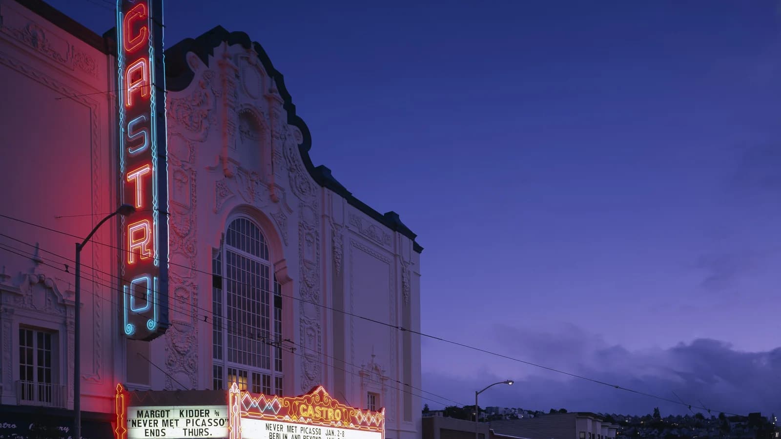 The tall neon Castro blade sign and lit marquee of the Castro Theatre's ornate white facade at dusk under a deep blue sky