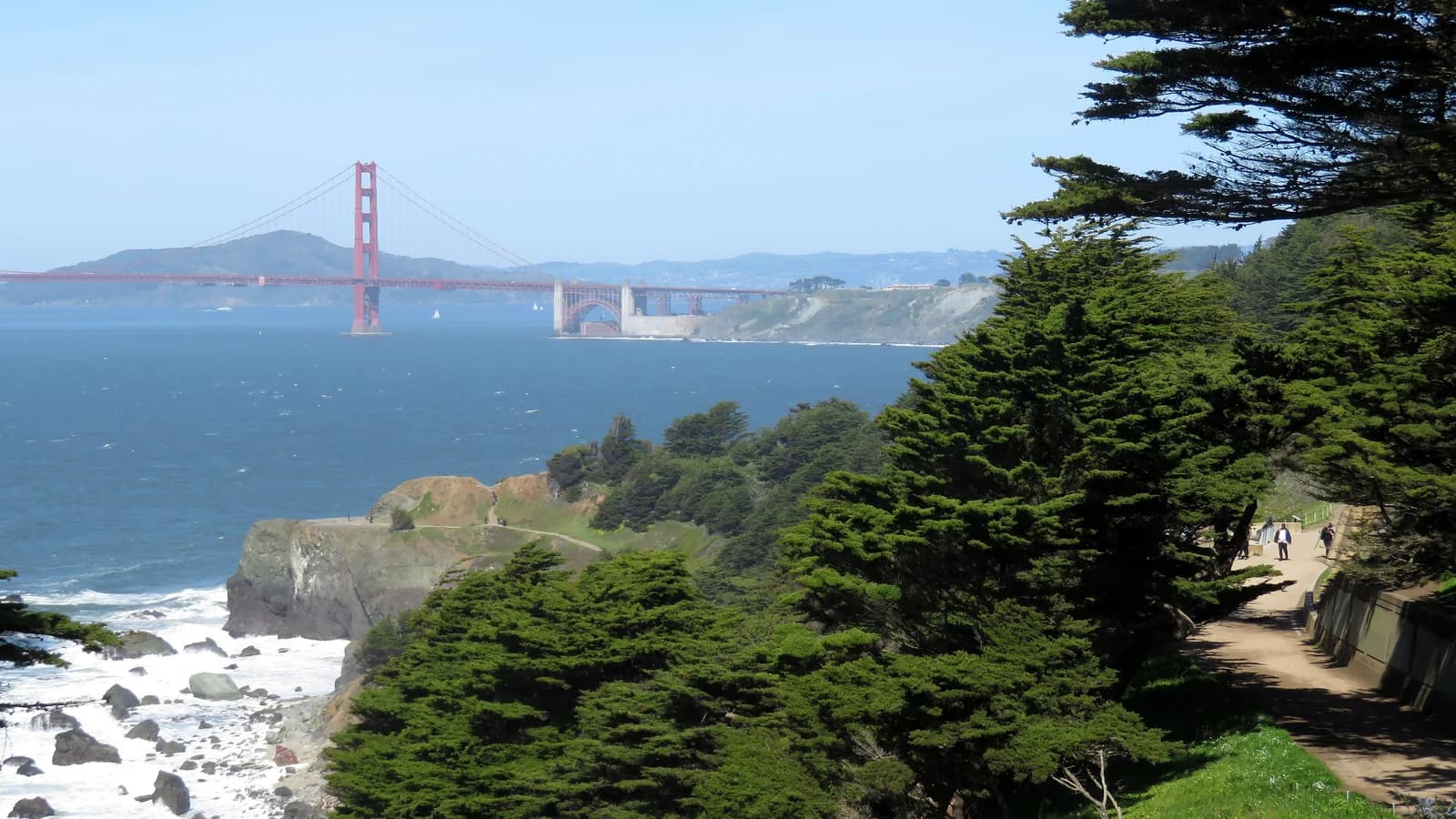 The Lands End Trail running past cypress trees above rocky cliffs and surf, with walkers on the path and the Golden Gate Bridge across the water