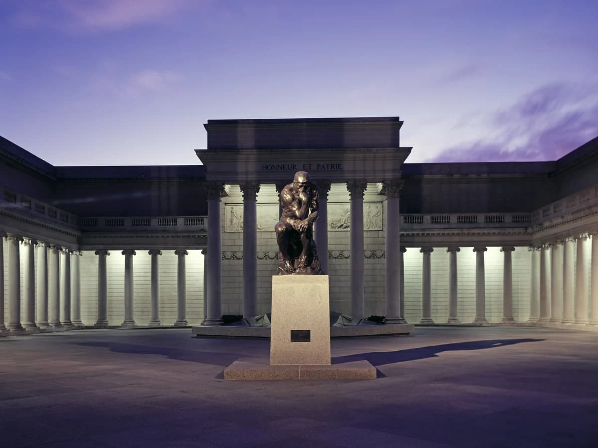 Bronze cast of Rodin's The Thinker on a stone pedestal in the Legion of Honor's courtyard, with lit colonnades and the central portico behind under a dusk sky