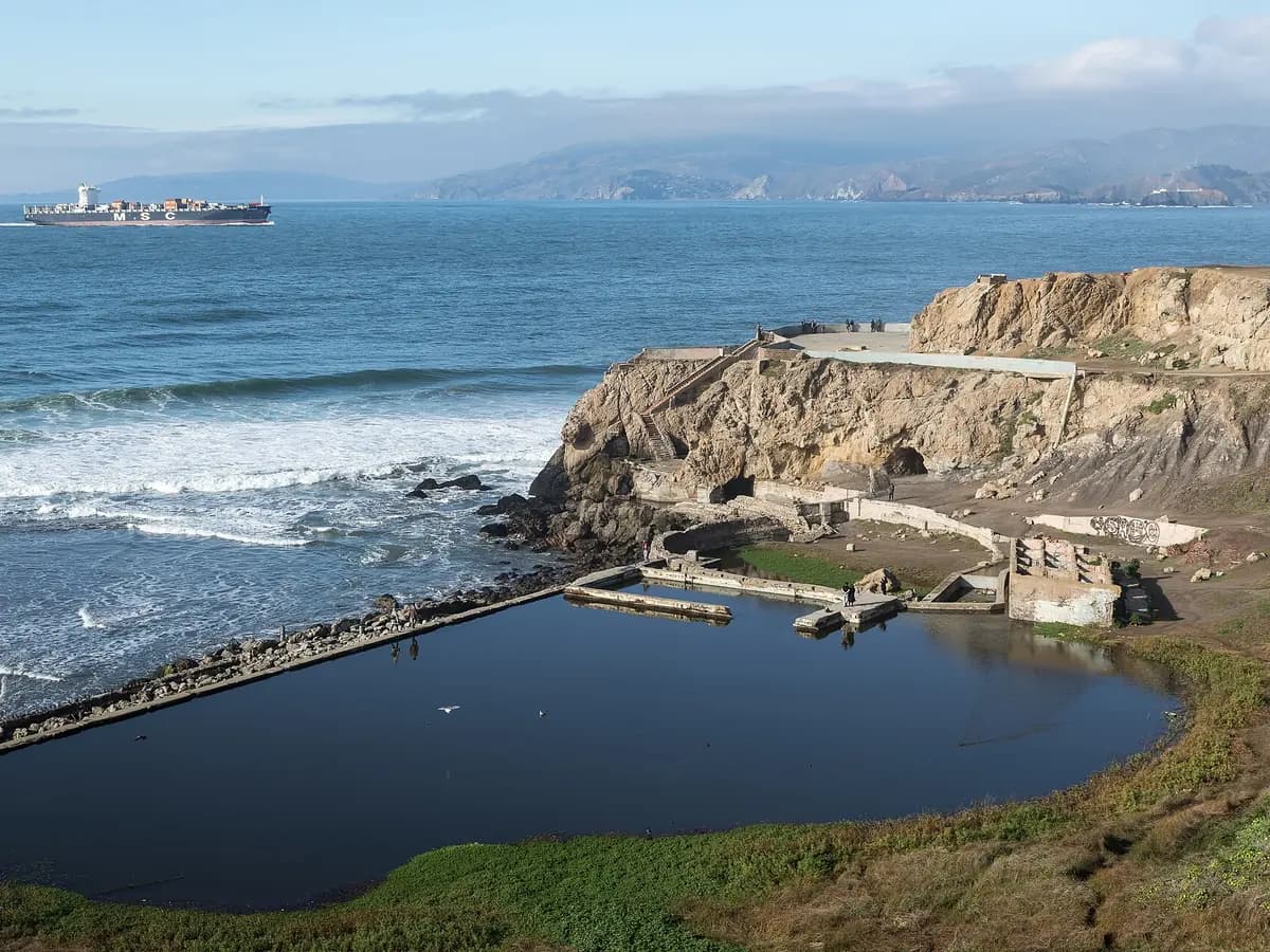 Concrete ruins and the flooded basin of the old Sutro Baths below a rocky headland with a cave tunnel, waves breaking offshore and a ship at sea