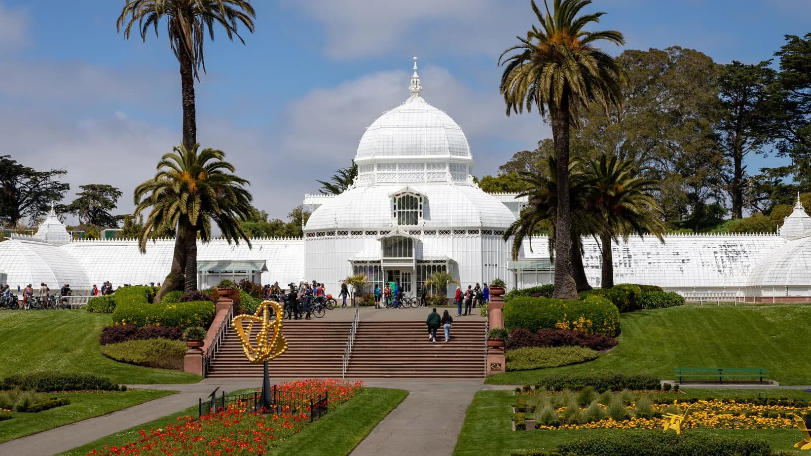 The white wood and glass Conservatory of Flowers with its central dome and palm trees, fronted by stairs, lawns and flower beds in Golden Gate Park