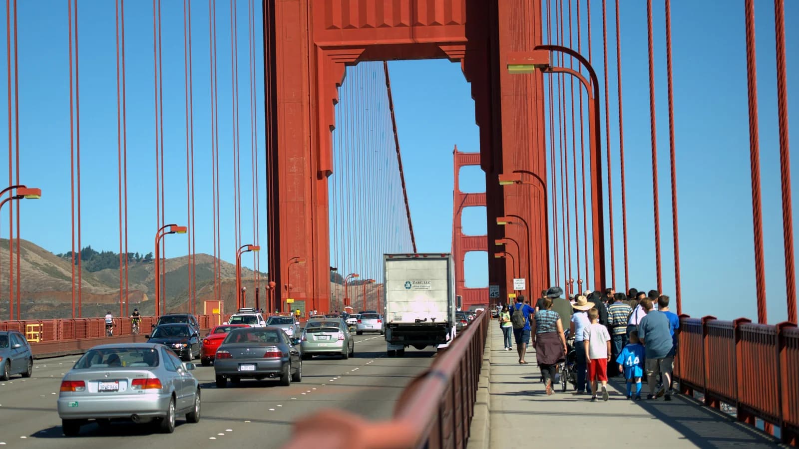 Walkers heading north on the Golden Gate Bridge's east sidewalk toward the orange south tower, with traffic alongside and Marin's hills beyond