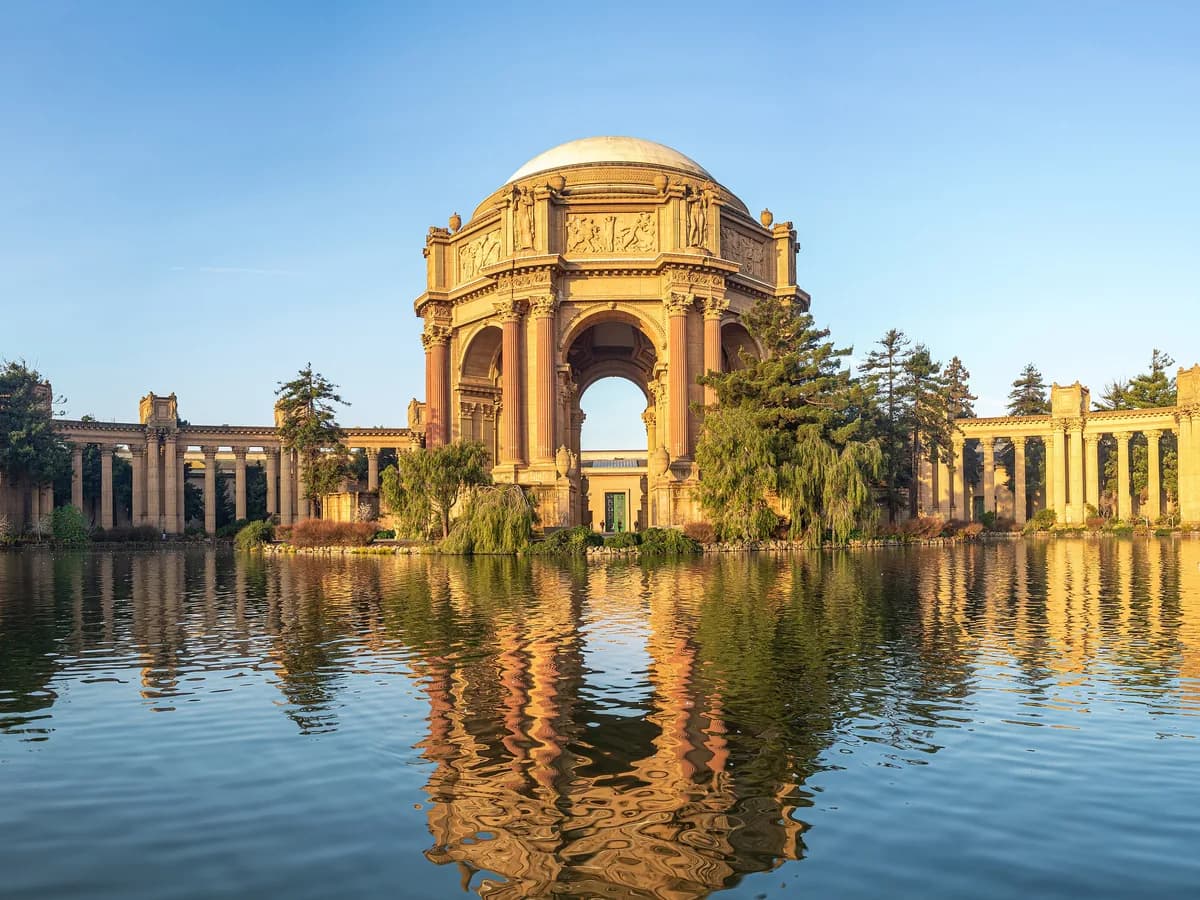 The domed rotunda and curving colonnades of the Palace of Fine Arts in warm low sun, reflected in the still lagoon under a clear blue sky