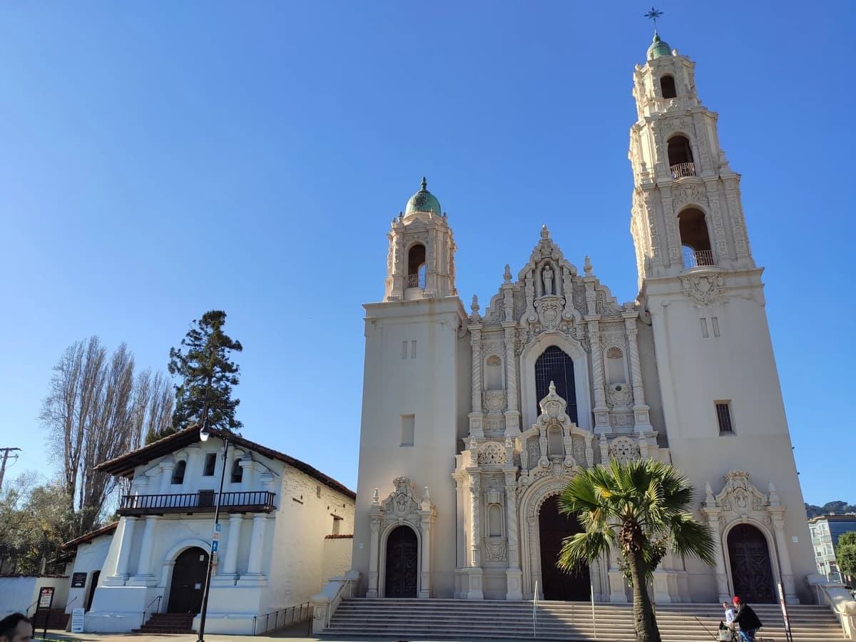 The small whitewashed Mission Dolores chapel with its tile roof and wooden balcony, beside the much taller ornate twin-towered basilica on Dolores Street, under a blue sky