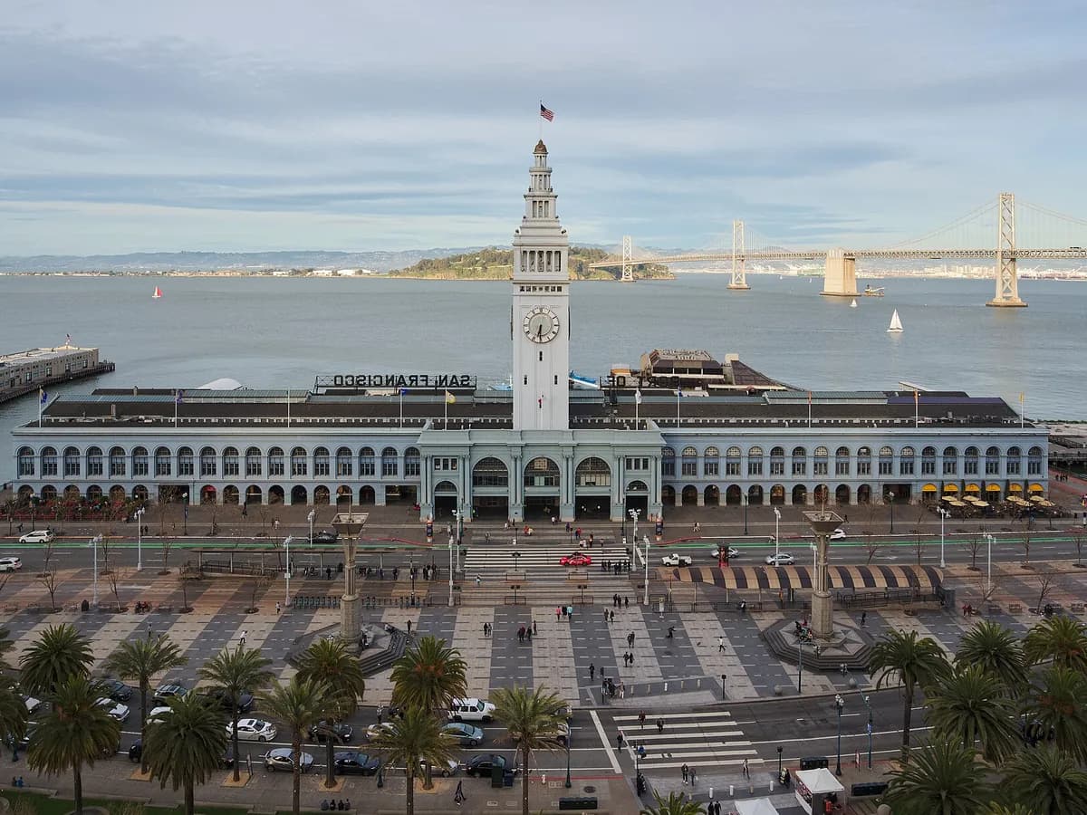 The Ferry Building's long arcaded facade and white clock tower on the Embarcadero, seen from above, with a palm-lined plaza in front and the Bay Bridge beyond