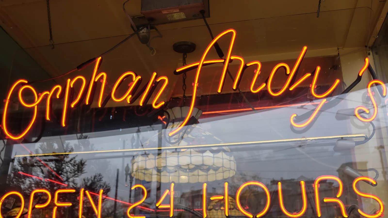 Orange neon script reading Orphan Andy's and Open 24 Hours in a window, with a Tiffany-style lamp hanging inside and the street reflected in the glass