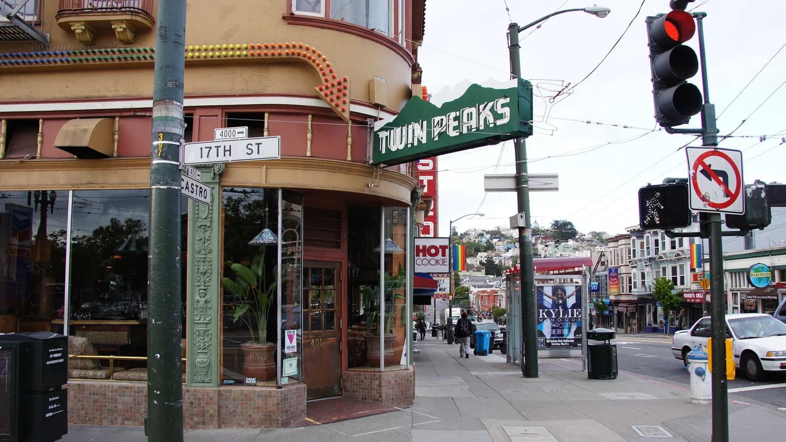 Twin Peaks Tavern's green sign over its corner plate-glass windows at Castro and 17th Street on an overcast day, with rainbow flags further down the street