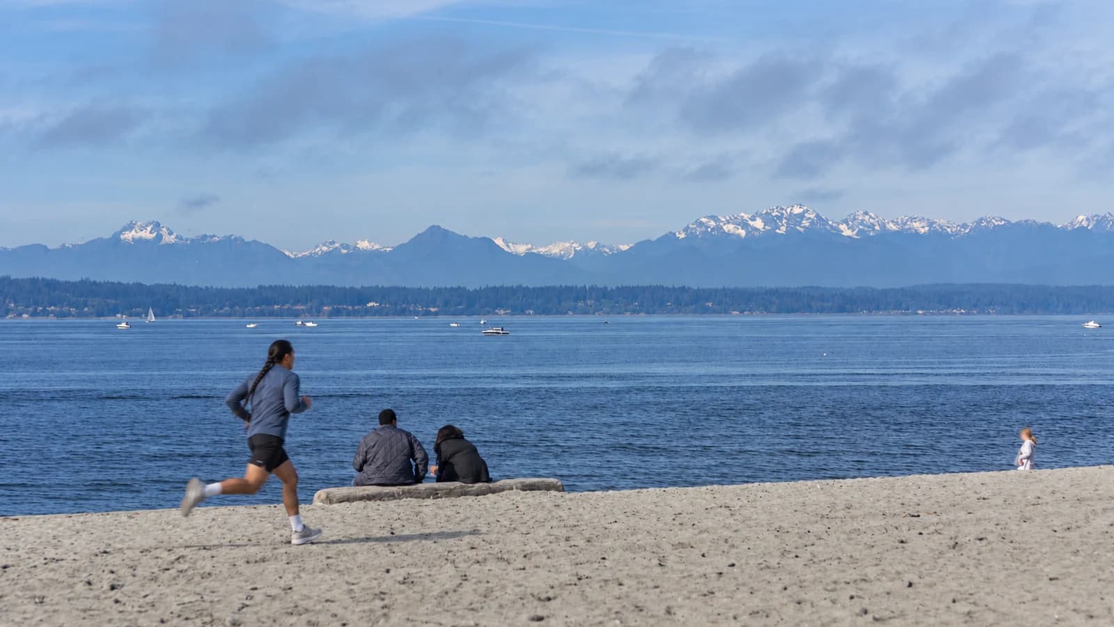 A runner on the sandy beach at Golden Gardens Park beside a driftwood log, with Puget Sound and the snow-capped Olympic Mountains on the horizon