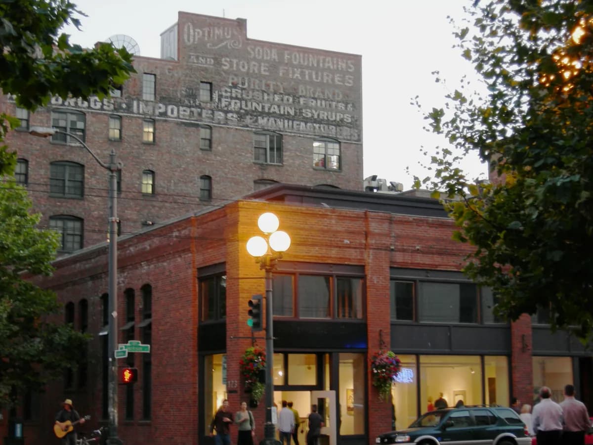 A red brick corner building with tall windows and an old painted sign on the wall above, beside leafy trees in Pioneer Square