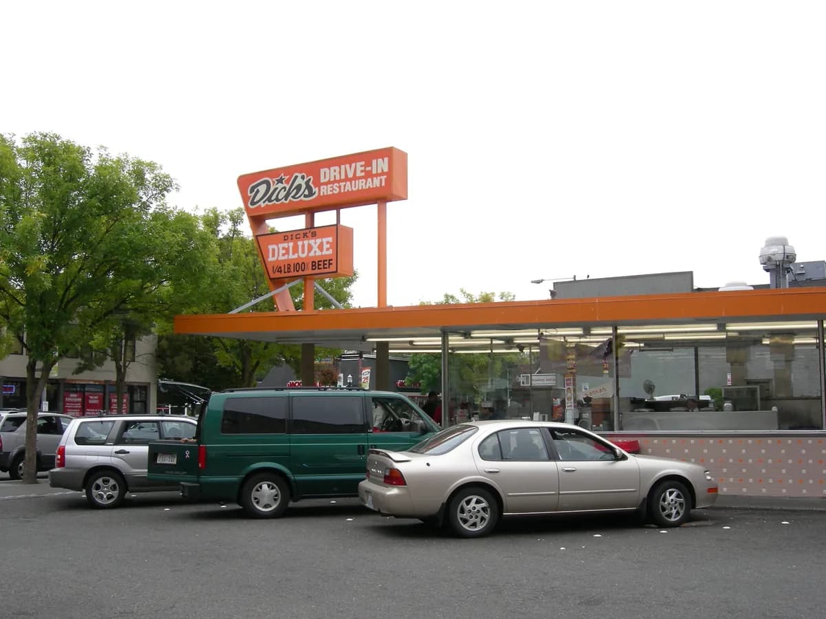 Dick's Drive-In on Broadway, a low orange roofed burger stand with a tall vintage sign, cars parked out front and trees behind