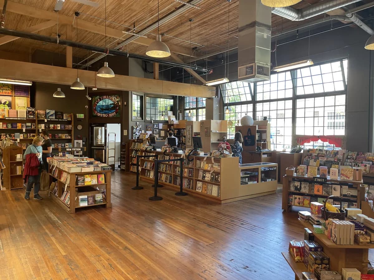 Inside Elliott Bay Book Company, wooden tables and shelves of books stand under exposed timber beams and tall industrial windows