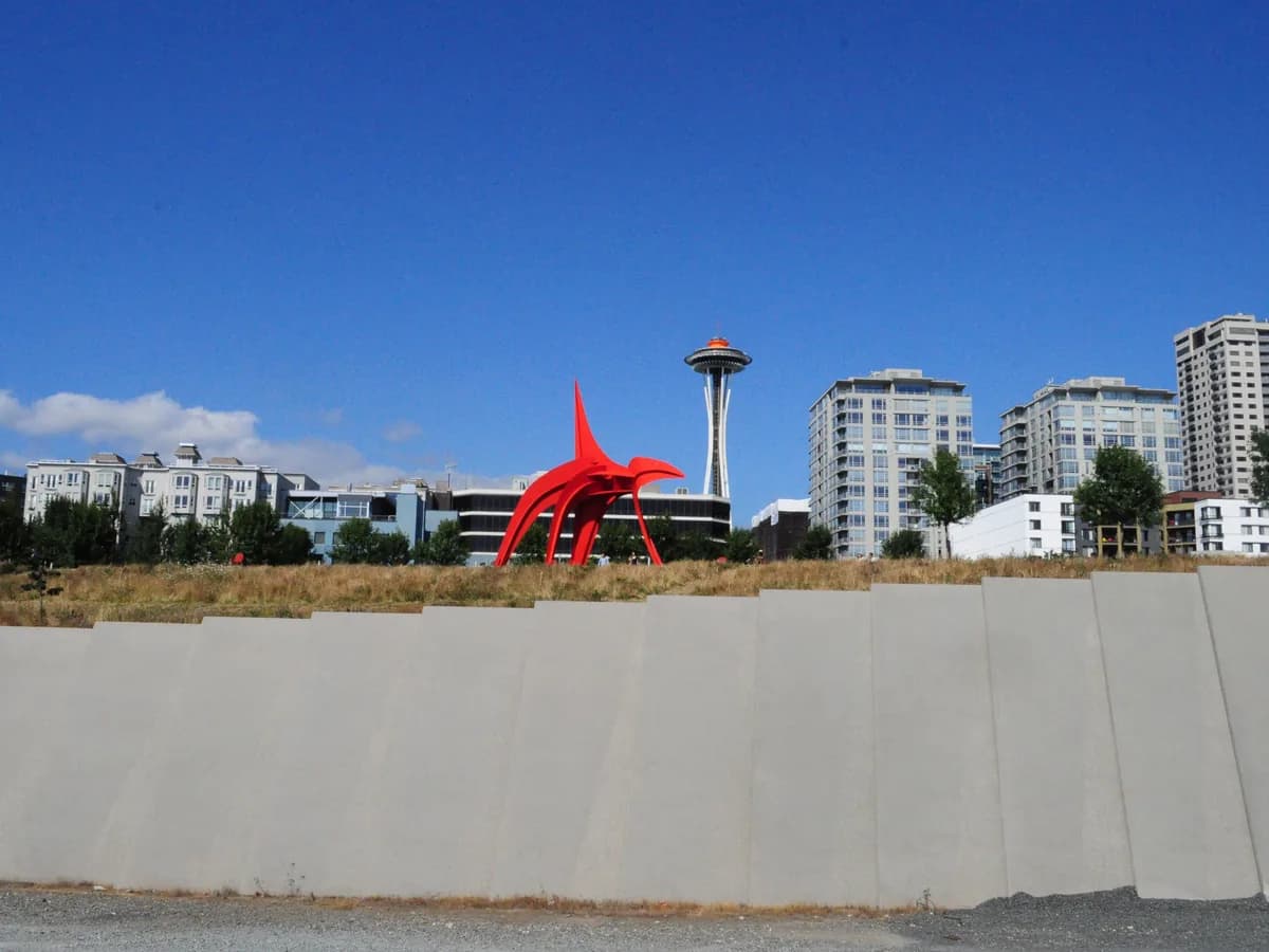 A huge red steel sculpture by Alexander Calder on a grassy terrace above a concrete wall, with the Space Needle and apartment towers behind under a blue sky