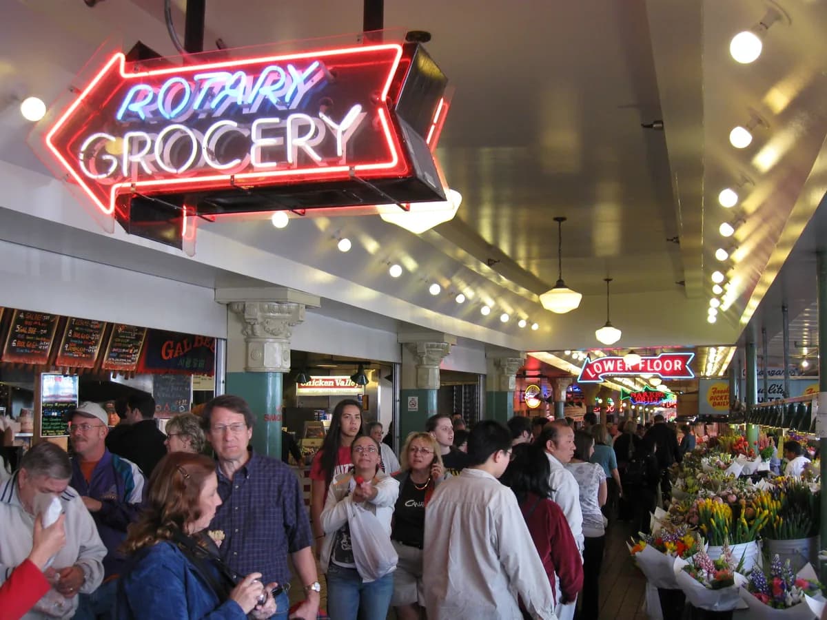 Shoppers crowd the covered Main Arcade of Pike Place Market under a red neon Rotary Grocery sign, with flower stalls and hanging lights
