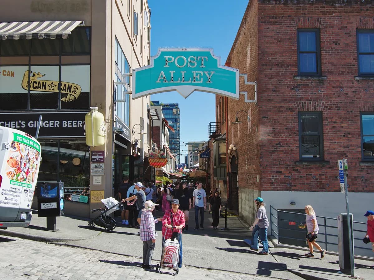 A green Post Alley sign hangs over a narrow brick lane off Pike Place Market, with shoppers walking under a clear blue sky