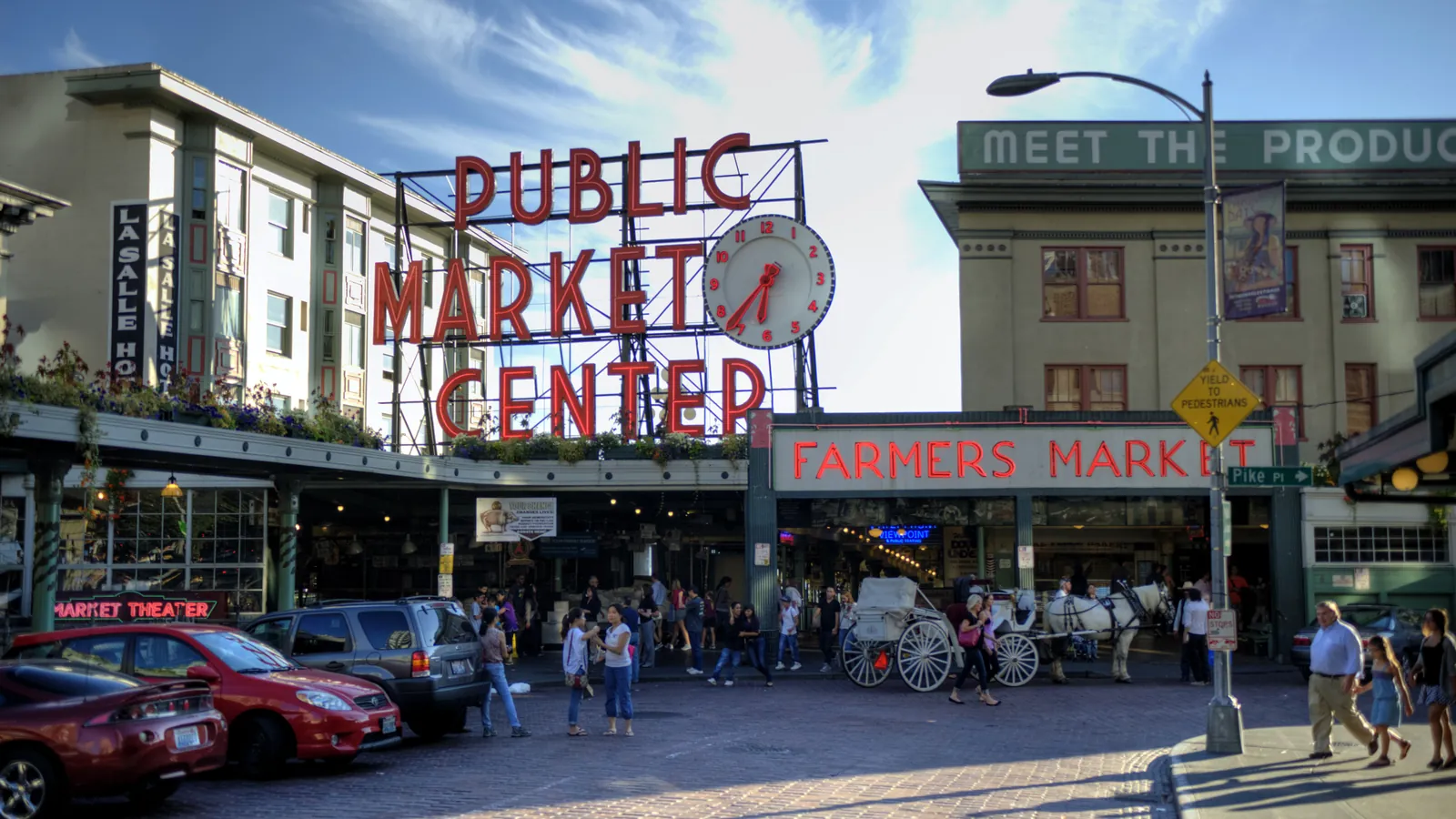 The red neon Public Market Center sign and clock above the entrance to Pike Place Market, with shoppers on the brick street below